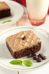 Chocolate bakery style cake brownie  with chocolate cream on top on a white plate and mint leaf and a glass of milk in the background