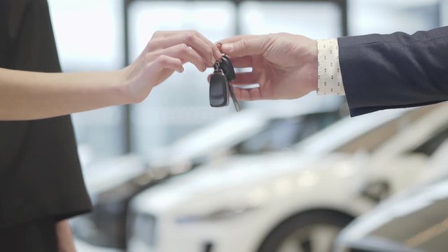 Unrecognized salesman in a business suit gives a car keys to unrecognized successful businesswoman and shake hands in the car dealership. Car showroom