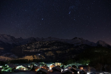 Winter mountain village in Hasliberg Hohfluh illuminated in a dark clear cold night with a sea of ​​stars and mountain range covered with snow in Switzerland, Europe. © Claudine