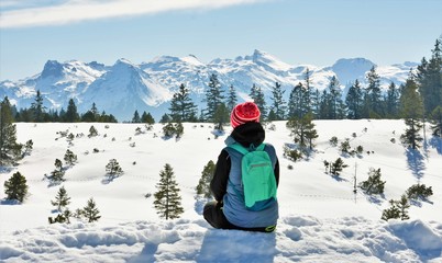 woman with a green backpack sits in the snow and looks into the distance at a beautiful view of the mountains