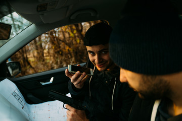 Two armed bandits sitting in a car planning their next robbery, while counting on a stopwatch the time they have for the robbery, showing off their guns and pointing at the blueprint.