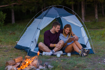 Young couple drinking coffee, enjoying the mountain view and checking the tablet with bonfire in the sunset