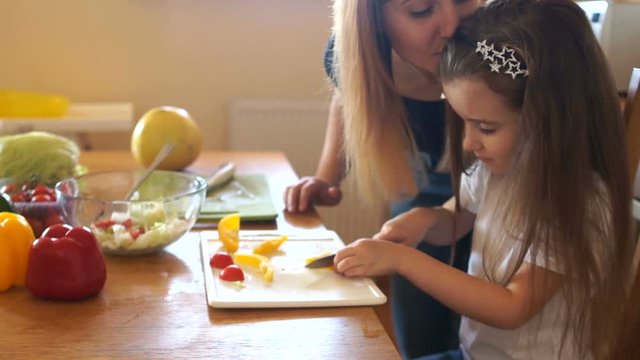 Loving mother kisses her daughter. A girl learns to cut vegetables for salad in the kitchen, her mother helps her