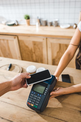 cropped view of cashier holding terminal wile man paying with smartphone in coffee house