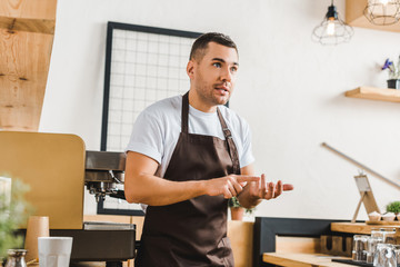 angry handsome barista in brown apron standing near coffee machine in coffee house