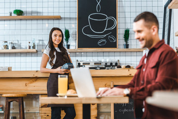 selective focus of waitress standing near bar counter with terminal wile freelancer in burgundy shirt sitting at table in coffee house