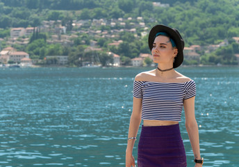 Fototapeta premium Traveler girl in a black hat and a striped T-shirt is sitting on the pier in the azure bay surrounded by mountains. Next to her is her backpack.