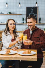 beautiful brunette woman and handsome man sitting at table with croissants and clinking glasses with juice in coffee house