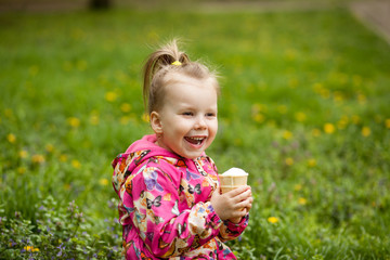 A small cute girl of 3-4 years old is sitting on the grass in early spring and eating a delicious ice cream in a waffle cup. The girl got dirty and dirty.