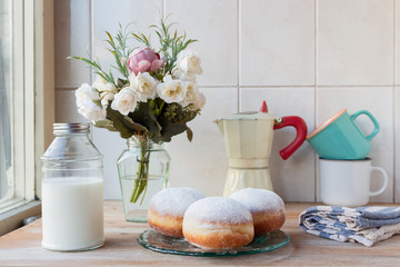 Three fresh doughnuts on a glass plate with coffee cups, moka pot, flowers and milk bottle in background, by the window