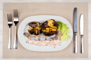 Baked potatoes and herring with sauce and salad on a white plate and cutlery.