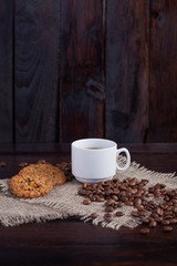 White cup of coffee with coffee grains near and biscuits on linen fabric on the dark vintage background