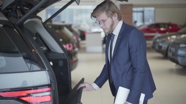 Bearded businessman choosing the car in motor show close up. Tall man holding tablet looks in opened trank and examines vehicle. Serious man choosing automobile in motor show. Concept of buying car