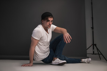 Fototapeta premium Young stylish guy posing sitting in the studio on a gray background. Dressed in a white T-shirt, blue jeans, a black hat and white sneakers. Model tests for men.