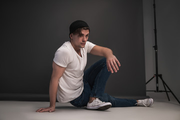 Young stylish guy posing sitting in the studio on a gray background. Dressed in a white T-shirt, blue jeans, a black hat and white sneakers. Model tests for men.