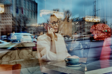 Attractive businesswoman in optical spectacles laughing during mobile conversation with friend via smartphone device.Positive young woman talking on telephone sitting at laptop computer in coffee shop