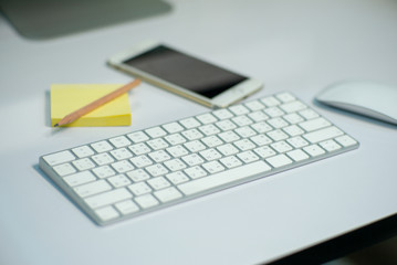 Selective focus on white keyborad on the desk with defocused stationary, mouse, and mobile phone in background