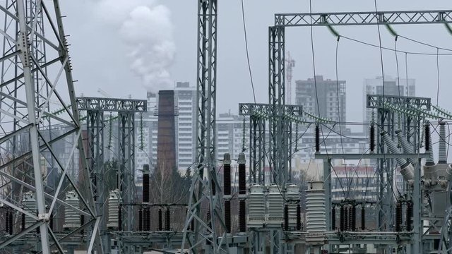 Gray-toned electric substation with a smoking pipe, transformers and power lines against the background of residential high-rise buildings.
