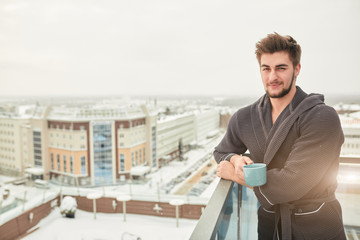 Cheerful adult man reaxing on terrace and wearing bathrobe. He is holding a cup of coffee in his...