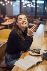 Gorgeous smiling young woman with dark hair eating cake and drinking coffee at a cafeteria in the evening sitting by the window. selective focus, noise effect