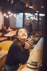 Gorgeous smiling young woman with dark hair eating cake and drinking coffee at a cafeteria in the evening sitting by the window. selective focus, noise effect