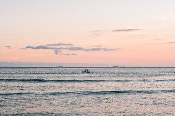 Sunset beach scenery with a boat leaving the beach on a colorful sky background and cargo ships...