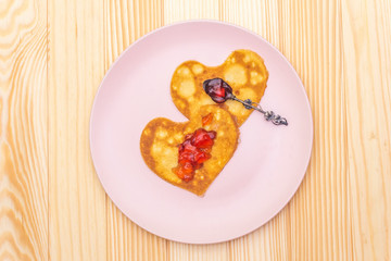 Heart shaped pancakes for romantic breakfast with strawberry jam and silver spoon. Shrovetide (carnival) concept. On wooden background, top view