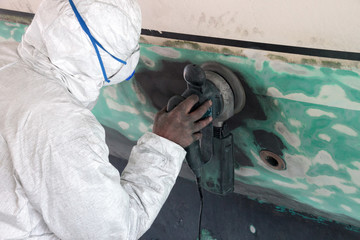 Worker wearing protection suit and mask is sanding down old paint from vessel