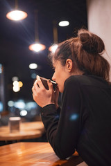 Gorgeous smiling young woman with dark hair eating cake and drinking coffee at a cafeteria in the evening sitting by the window. selective focus, noise effect