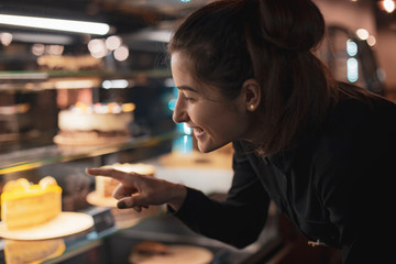 Gorgeous smiling young woman with dark hair eating cake and drinking coffee at a cafeteria in the evening sitting by the window. selective focus, noise effect
