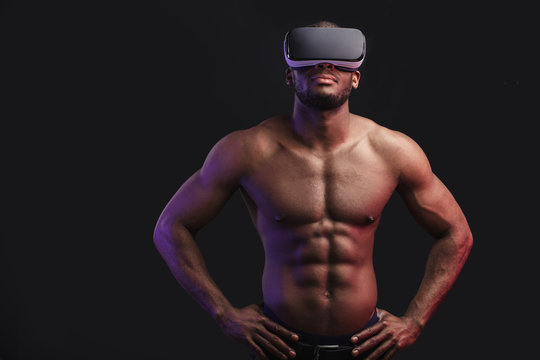 Strong African boxer with bare chest wearing mobile virtual reality headset prepares to fight with virtual opponent, standing isolayed against dark wall. Sport and augmented virtual reality concept