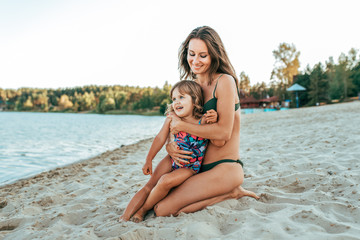 A young mother plays with her daughter girl of 2-5 years, on beach in summer, an outdoor resort by the lake. Happy rest on the weekend. Bright swimsuit on child. Parents love family and parenting.