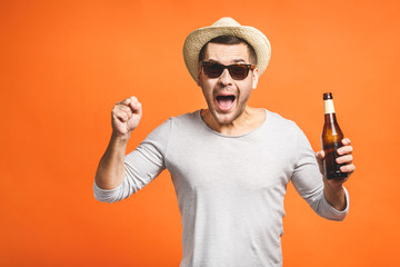 A young guy with a hat and sunglasses isolated on a orange background holds a bottle of beer....