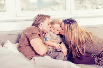 Portrait of happy family sitting on the sofa and playing. Parents kissing their baby son.