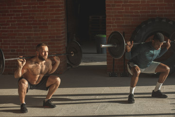 Obraz na płótnie Canvas Sport, crossfit lifestyle and people concept - two determined caucasian and african male athletes with barbell lifting weight at indoor workout against brick walls