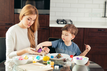 Blonde Female babysitter having confidence, skill and understanding to support children's play, interacting with boy in a playful way, painting eggs to Easter celebration.