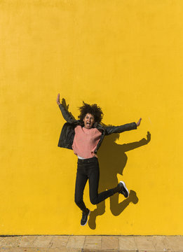 Black woman with afro hair jumping for joy in the street with a yellow wall in the background
