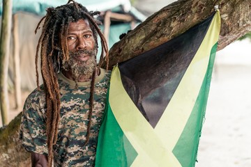 African American bearded male with dreadlocks holding Jamaica flag near tree