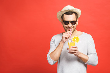 Portrait of attractive young man in hat and sunglasses standing and drinking orange juice over red...