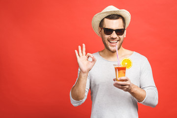 Portrait of attractive young man in hat and sunglasses standing and drinking orange juice over red...