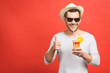 Portrait of attractive young man in hat and sunglasses standing and drinking orange juice over red...
