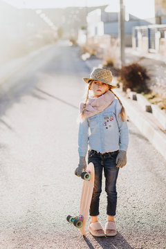 Cute little girl in trendy outfit holding skateboard and looking away while standing on road on sunny day on suburban street