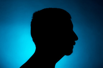 Cleanly defined silhouette of a male person turned to the left against a blue background with a spotlight and bright area right behind the bust. Studio shot with well defined colour background.