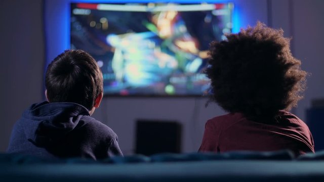 Rear view teenager boys with joysticks fighting shooter game on home console, looking at blurry wide screen plazma TV. Two best friends eating during game sitting on sofa in domestic room at night