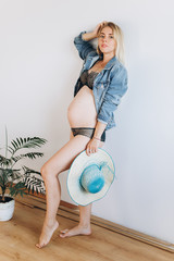 Pregnant cheerful happy future mother expecting for a baby and posing indoors on white background in fashion lingerie and jeans coat with hat. Pregnancy, beauty, fashion concept