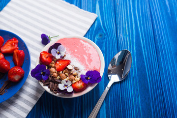 Pink yogurt bowl with strawberry, nuts, chocolate, decorated with flowers, with spoon on navy wooden background, top view.