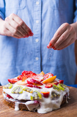 Female hands sprinkled sour cream fruit cake with coconut flakes at home.