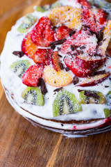 Closeup homemade sour cream fruit cake with coconut and chocolate flakes, kiwi,strawberry and peach on wooden table.