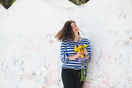Summer young woman in stripped vest with vibrant bouquet of yellow freesias and irises near light concrete wall.