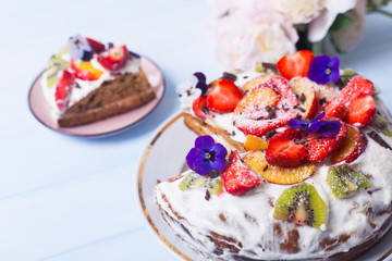 Closeup spring homemade sour cream fruit cake with coconut and chocolate flakes, kiwi,strawberry and peach, decorated with flowers with cut piece on wooden table.